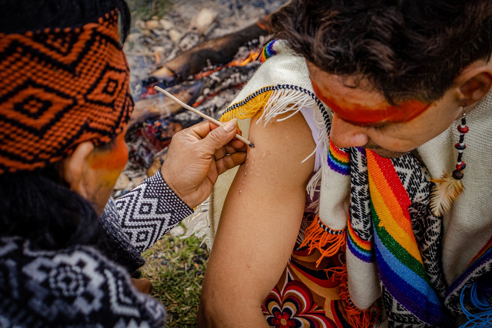 Traditional Kambo ceremony with a practitioner dressed in colorful textiles, face paint, beads, and feathers, carefully applying Kambo medicine to a participant's arm.
