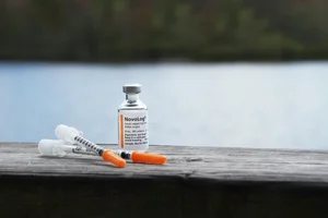 A vial of NovoLog insulin and two syringes on a wooden surface near water