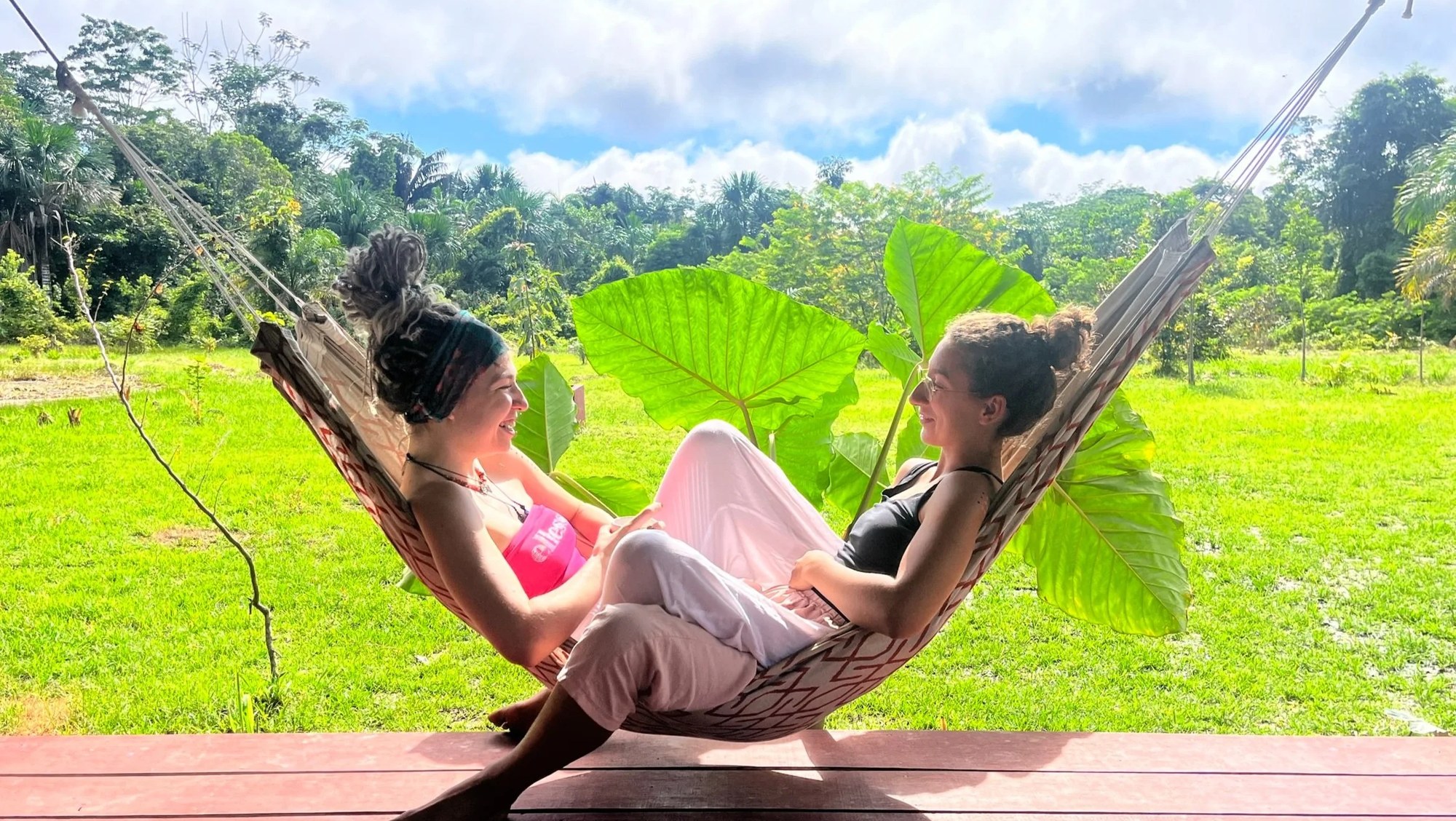 Two people relaxing in hammocks on a wooden porch overlooking green fields and jungle in the Amazon.