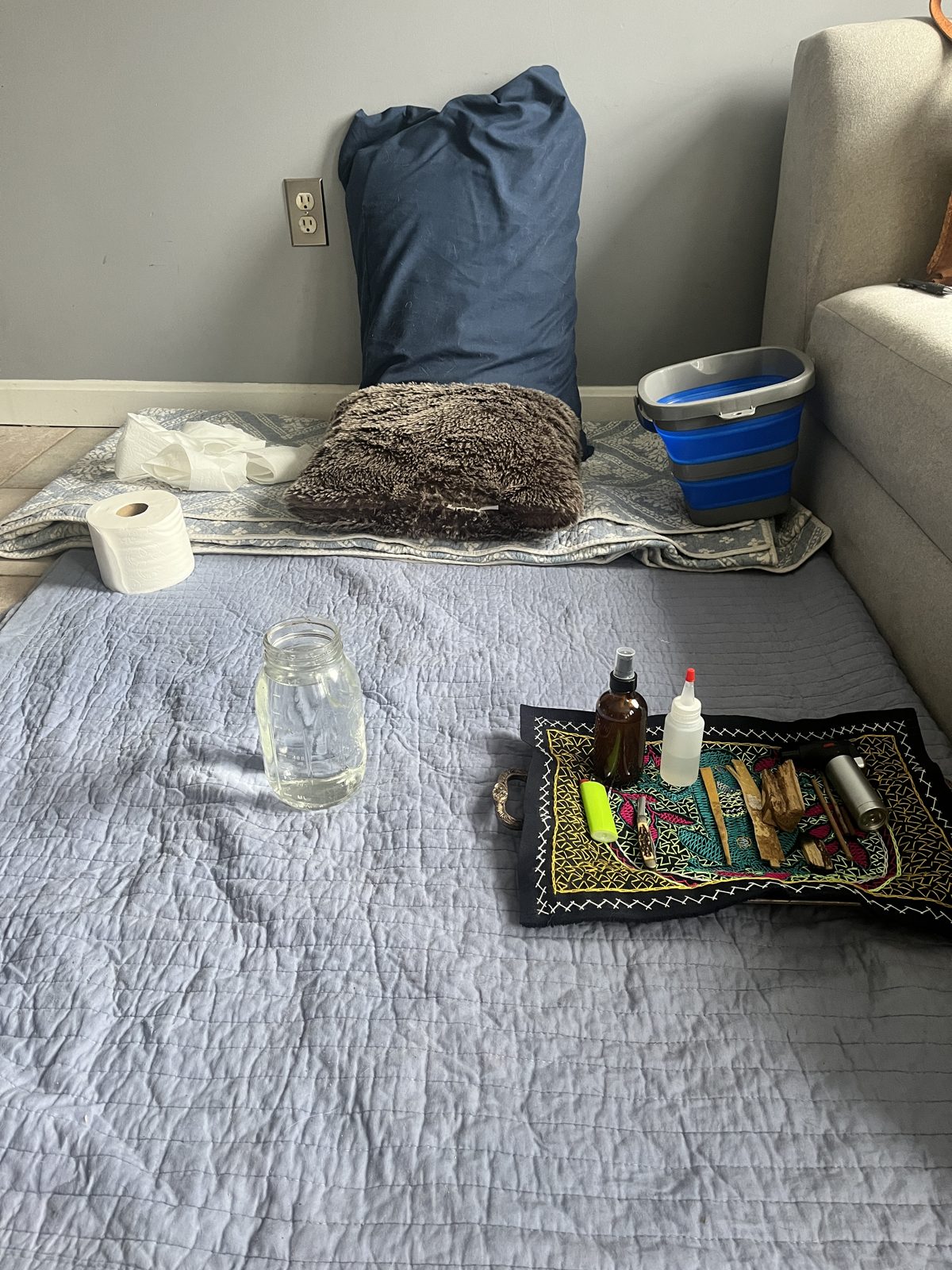 Indoor Kambo ceremony setup featuring a blanket on the floor, pillows for comfort, a blue collapsible bucket, toilet paper, a mason jar with liquid, and a tray with bottles and small ceremonial items.
